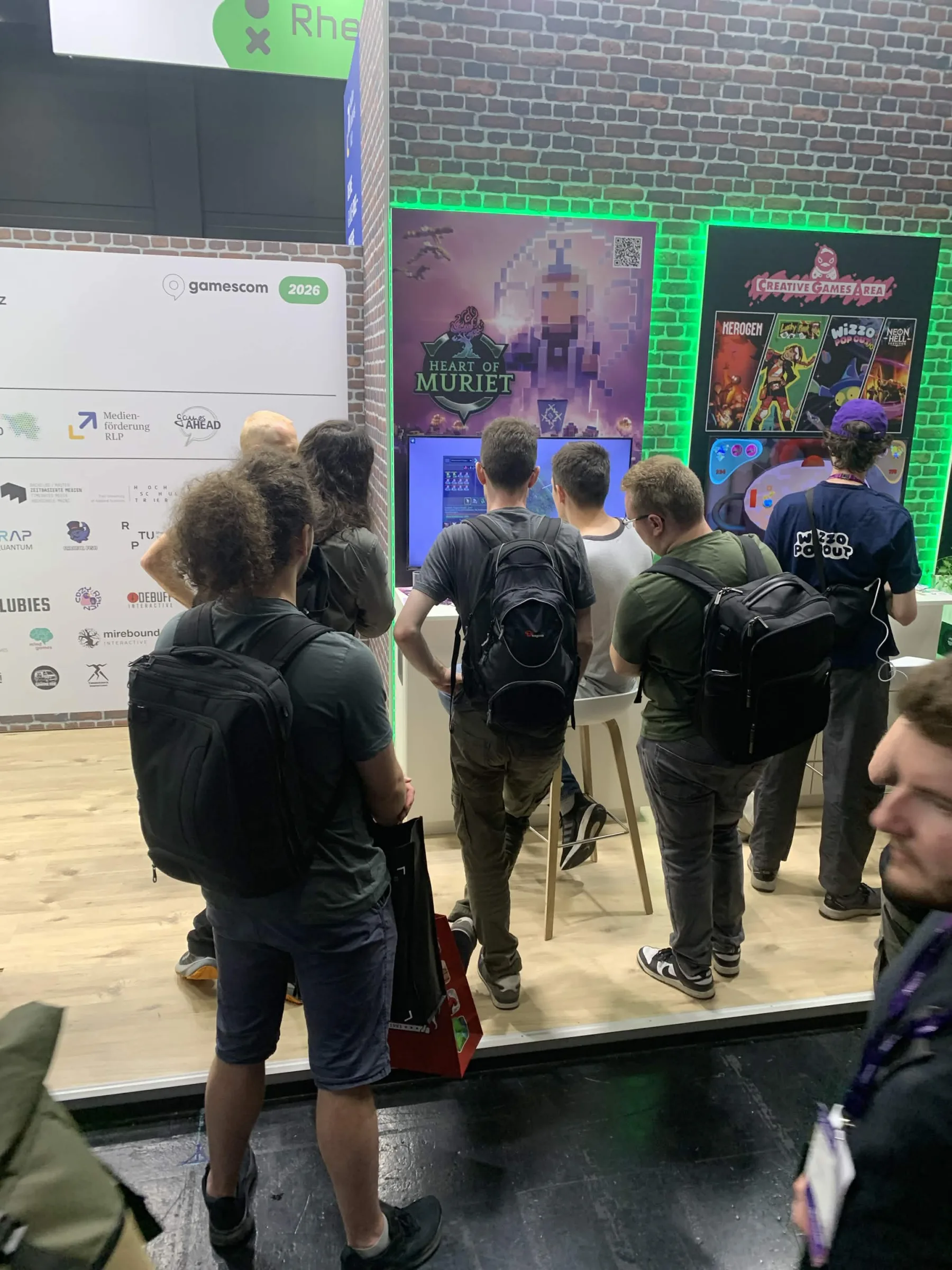 A crowd gathered around the Heart of Muriet demo station under green booth lighting at gamescom